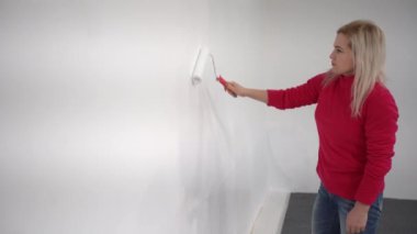 Kapatın. Yeni evde tek başına resim yapan oldukça neşeli beyaz bir kadının yan görüntüsü. Güzel bir kadın evi yeniliyor. Duvarı paten fırçasıyla boyuyor. Daireyi yeniden tasarlıyor.