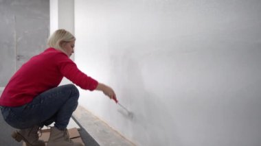 Kapatın. Yeni evde tek başına resim yapan oldukça neşeli beyaz bir kadının yan görüntüsü. Güzel bir kadın evi yeniliyor. Duvarı paten fırçasıyla boyuyor. Daireyi yeniden tasarlıyor.