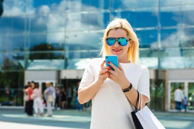 Güzel kız alışveriş torbalarını tutuyor, akıllı bir telefon kullanıyor ve şehirde alışveriş yaparken gülümsüyor.