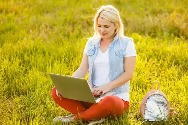 Mladá žena s laptopem sedí na trávě v parku za slunečného dne. Smějící se blondýna v bílém tílku a šortkách. Online školení, vzdálená práce a komunikace v sociálních sítích — Stock fotografie