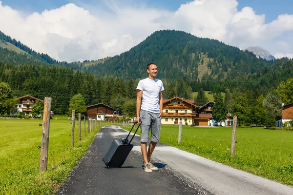 Yolcu adam elinde bavulla dağlarda sokakta yürüyor. Seyahat kavramı. — Stok fotoğraf