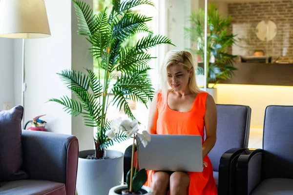 Une photo vue de côté de la femme utilisant un ordinateur portable pour le e-shopping tout en étant assis dans un hall d'hôtel moderne. Une femme blonde portant une robe regarde l'écran — Photo