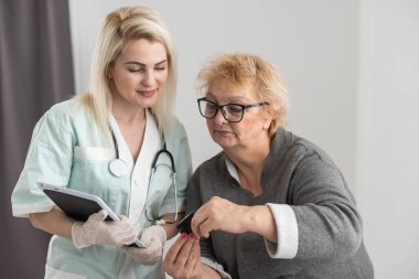Parmak ucu nabız oksimetresi olan kadın bakıcı, yaşlı, sağlık kontrolüne bağlı, COVID-19 veya Coronavirus salgını sırasında evdeki oksijen doygunluğunu ve kalp atışlarının hızını ölçüyor..