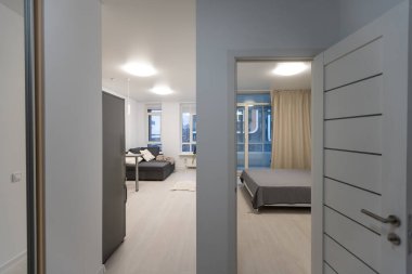 Bir sürü beyaz kapısı ve siyah kapı kolu olan küçük ve beyaz apartman koridoru, ahşap zemin ve yatak odası kapıları açık.