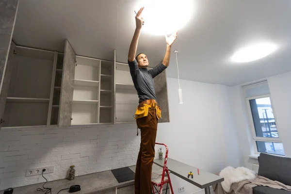 Ηλεκτρολόγος, ένας άνδρας ηλεκτρολόγος στέκεται στις σκάλες κρατώντας καλωδιώσεις στα χέρια του και γδύνοντας, επισκευάζοντας το φως στο σπίτι, επισκευάζοντας τις εργασίες, καλέστε τον πλοίαρχο, ηλεκτρολόγος άνθρωπος επισκευάζοντας το φως — Φωτογραφία Αρχείου
