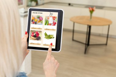 Mutfakta dijital tablet kullanan bir kadın çevrimiçi yiyecek alışverişi yapıyor, tablet ekranda yakın çekim yapıyor. Mobil aygıtlar kullanılarak çevrimiçi satın alma kavramı.