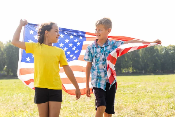 Счастливая кавказская девочка и мальчик улыбаются, держа за руки и размахивая американским флагом на улице, празднуя 4 июля, День независимости, День флага концепция. — стоковое фото