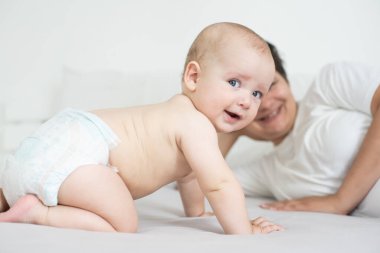 Anne ve çocuk beyaz bir yatakta. Anne ve bebek bezli çocuk yatak odasında oynuyorlar. Ebeveyn ve küçük çocuk evde dinleniyor. Aile birlikte eğleniyor..