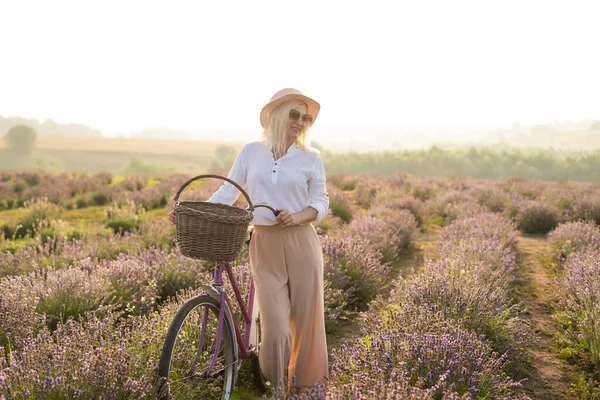 Jovem mulher loira viajante usando chapéu de palha no campo de lavanda cercada com flores de lavanda. — Fotografia de Stock