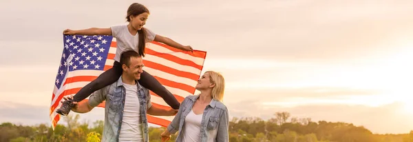 Hermosa familia con la bandera americana en un campo al atardecer. Día de la Independencia, 4 de julio. — Foto de Stock