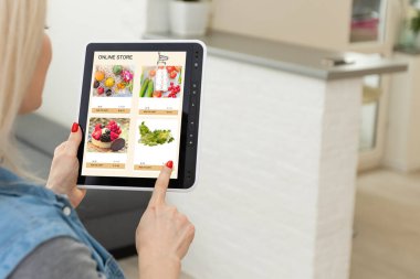 Mutfakta dijital tablet kullanan bir kadın çevrimiçi yiyecek alışverişi yapıyor, tablet ekranda yakın çekim yapıyor. Mobil aygıtlar kullanılarak çevrimiçi satın alma kavramı.
