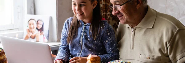 Grandfather and granddaughter are talking via video link to their friends. Decorated table with colorful eggs and cake. Chatting during the COVID pandemic and the Easter holidays — Stock Photo, Image