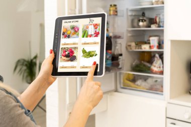 Mutfakta dijital tablet kullanan bir kadın çevrimiçi yiyecek alışverişi yapıyor, tablet ekranda yakın çekim yapıyor. Mobil aygıtlar kullanılarak çevrimiçi satın alma kavramı.