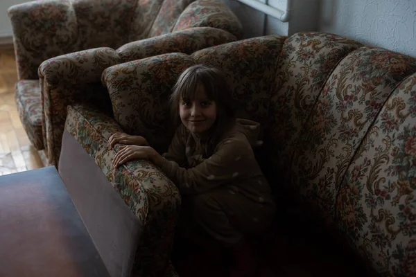 Una niña triste escondida detrás de un sofá en casa. Concepto de violencia doméstica —  Fotos de Stock