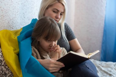 Anne ve kızı yatakta Ukrayna bayrağıyla kitap okuyorlar.