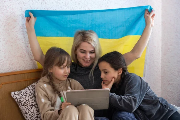 Familien gegen den Krieg. Mutter und zwei Töchter lesen mit der Fahne der Ukraine im Bett. — Stockfoto