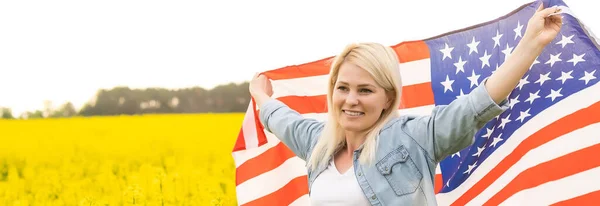 Mujer atractiva sosteniendo una bandera americana en el viento en un campo de colza. Paisaje de verano contra el cielo azul. Orientación horizontal. — Foto de Stock