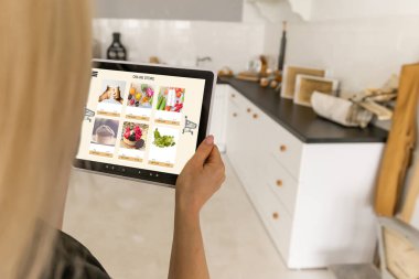 Mutfakta dijital tablet kullanan bir kadın çevrimiçi yiyecek alışverişi yapıyor, tablet ekranda yakın çekim yapıyor. Mobil aygıtlar kullanılarak çevrimiçi satın alma kavramı.