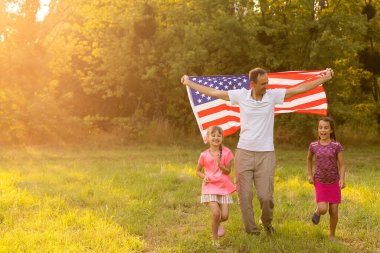 Arka bahçelerinde birlikte oturan mutlu aile arkalarında Amerikan bayrağını taşıyor. Amerika 'nın bağımsızlık gününü çocuklarıyla kutlayan gülümseyen çift Amerikan bayrağı taşıyor.
