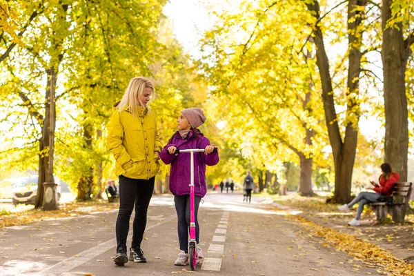 Youg moeder wandelen met haar dochter rijden een peuter scooter in park. — Stockfoto