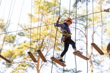 Küçük kız bir ipin üzerinde duruyor, elleriyle bir ip tutuyor. İp parkındaki bir çocuk engelleri aşıyor