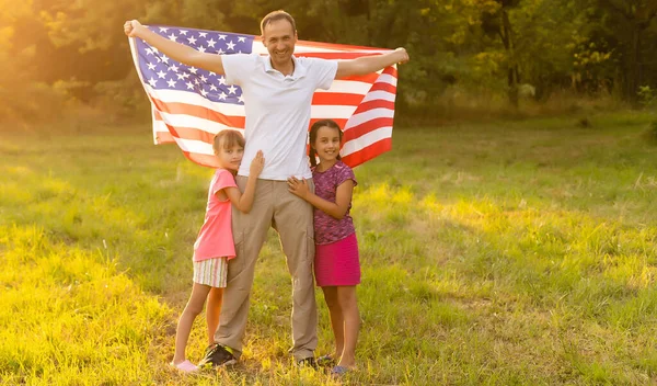 Lycklig familj sitter tillsammans på sin bakgård och håller den amerikanska flaggan bakom sig. Leende par med sina barn firar amerikanska självständighetsdagen hålla amerikansk flagga — Stockfoto