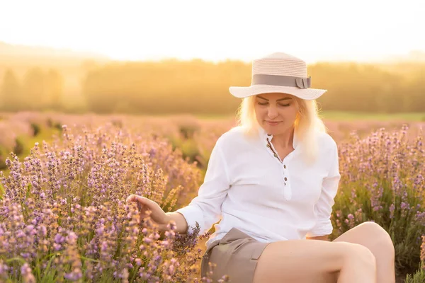 Piękna młoda zdrowa kobieta w białej sukience biegnąca radośnie przez pole lawendy trzymając słomkowy kapelusz pod promieniami zachodzącego słońca — Zdjęcie stockowe