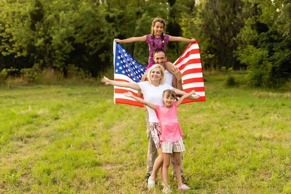 Happy family sitting together in their backyard holding the american flag behind them. Smiling couple with their kids celebrating american independence day holding american flag — Stock Photo, Image