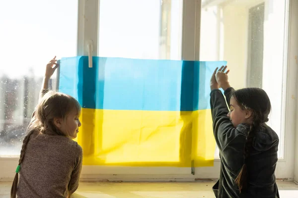 Kleine Mädchen mit ukrainischer Flagge vor einem Fenster. Die kleinen Mädchen schwenken die Nationalflagge, während sie für den Frieden beten — Stockfoto