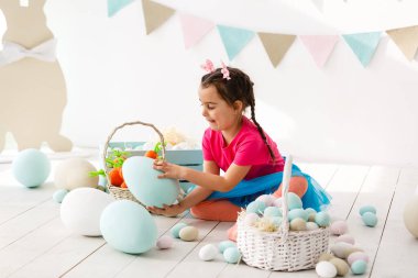 Paskalya 'ya hazırlanıyorum. Paskalya yumurtası tutan ve arka planda süslemelerle gülümseyen sevimli küçük kız.