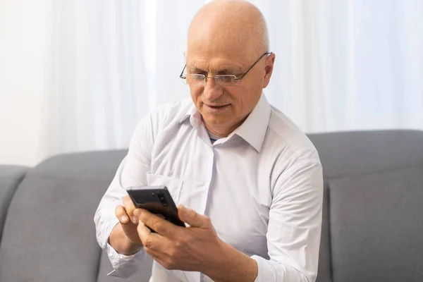 Uomo anziano chatta su un telefono cellulare mentre si rilassa su un divano nel suo soggiorno — Foto Stock