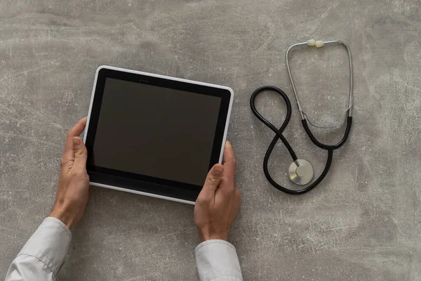 Доктор рука тримає планшет з порожнім екраном і стетоскопом на сірому столі, копіювати простір, вид зверху — стокове фото