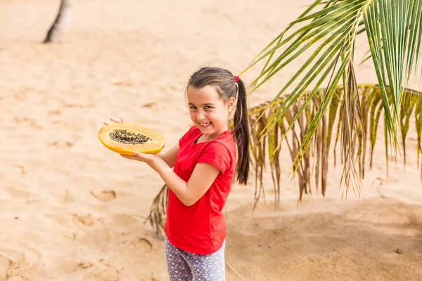После игры на море черный песок пляжа маленькая милая девочка пикники натуральной вегетарианской пищи - есть апельсиновый кусок свежей спелых папайи, здоровый образ жизни детей в отпуске на тропическом острове — стоковое фото