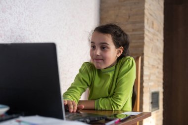 Yüzünde kulaklıkla gülümseyen küçük beyaz kız bilgisayarını kullanarak uzaktan öğretmeniyle video görüşmesi yapıyor, öğretmeniyle mutlu küçük çocuk dalgası selamlaması yapıyor, bilgisayarda çalışıyor, evde eğitim kavramı geliştiriyor.