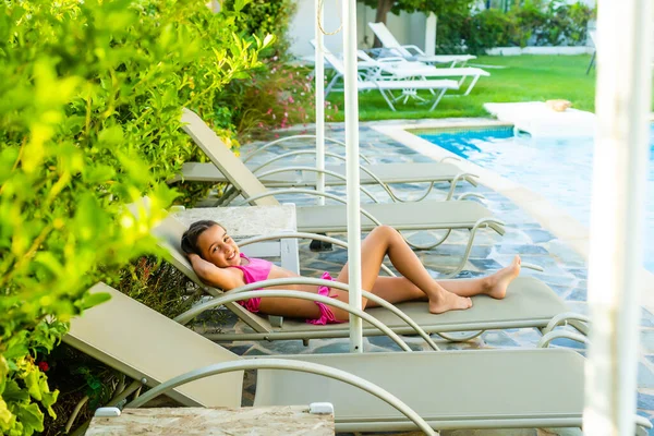 Belle petite fille reposant sur une chaise longue à côté de la piscine sur fond de rangées de chaises longues, pour se reposer ou bronzer. — Photo