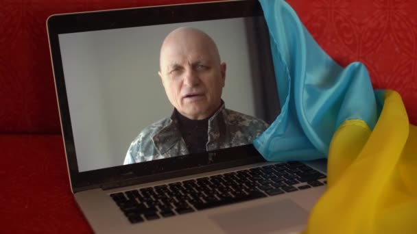 Στρατιώτης με στρατιωτική στολή κάθεται στον καναπέ στο σπίτι χρησιμοποιώντας φορητό υπολογιστή webcam για την επικοινωνία με την οικογένεια. Ουκρανική σημαία στο παρασκήνιο. Στρατιωτικές ενέργειες στην Ουκρανία. Ελεύθερος δημοκρατικός λαός στο κράτος — Αρχείο Βίντεο