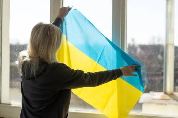 Mulher embrulhada na bandeira da Ucrânia olhando pela janela. espaço de cópia — Fotografia de Stock