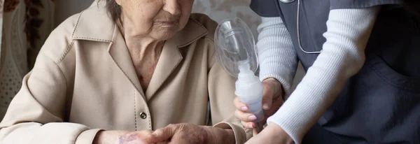 Young high-skilled pleasant woman doctor putting on nebulizer mask on face of her elderly female patient to make inhalation when visitng him at home. Flu, cold and cough treatment. — Stock Photo, Image