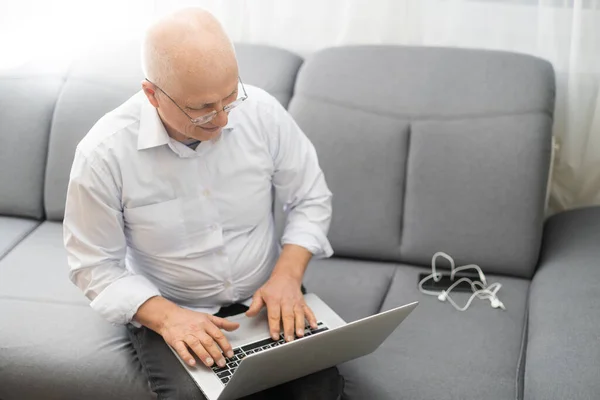 Elderly bald man in white shirt and black pants correcting glasses and working on laptop while sitting in living room. — Stock Photo, Image