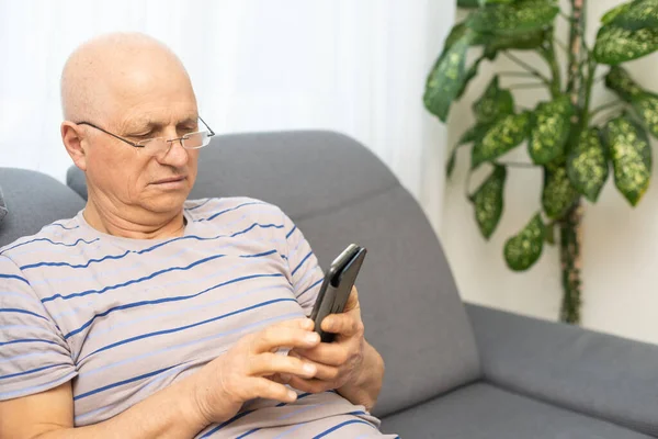 Uomo anziano chatta su un telefono cellulare mentre si rilassa su un divano nel suo soggiorno — Foto Stock