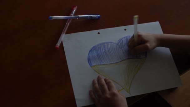 Las manos de un niño dibujan un icono en forma de corazón con la imagen de la Bandera Nacional de Ucrania. Vista desde arriba. Niños contra la guerra. Niños dibujando por la paz en Ucrania — Vídeos de Stock