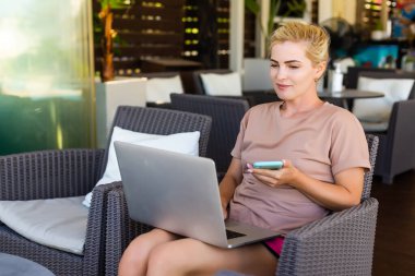 Genç bir bayan, halka açık kablosuz internet alanında dizüstü bilgisayarda çalışıyor, yazı yazıyor, insanlar arka planda geçip gidiyor.