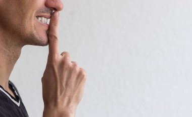 Sır olarak sakla. Tıraşsız, beyaz tişörtlü genç adam sessizlik jesti yapıyor, sessiz duruyor ve gizemli bir yüz ifadesiyle kameraya bakıyor.