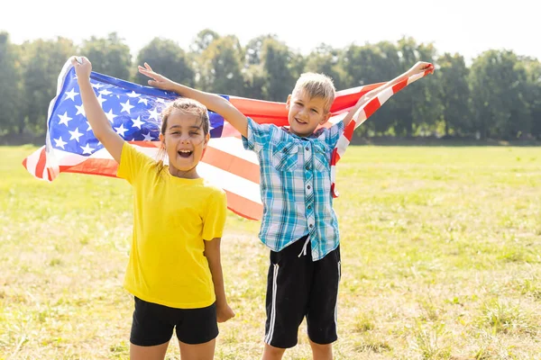 Счастливая кавказская девочка и мальчик улыбаются, держа за руки и размахивая американским флагом на улице, празднуя 4 июля, День независимости, День флага концепция. — стоковое фото