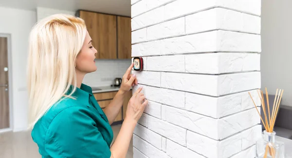 Женщина регулирует температуру отопления с помощью современного беспроводного термостата, установленного на белой стене дома. Концепция интеллектуального отопления — стоковое фото