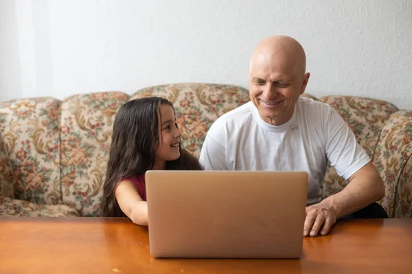 Adorable petite fille étreignant heureux grand-père en utilisant un ordinateur portable à la maison . — Photo