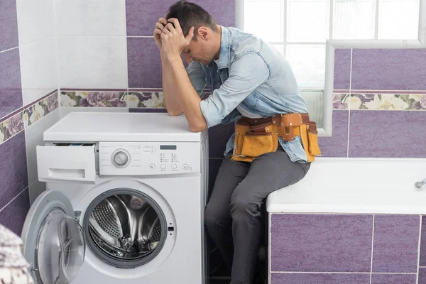 O jovem e bonito reparador em terno de trabalhador com a caixa de ferramentas profissionais está consertando a máquina de lavar roupa no banheiro — Fotografia de Stock