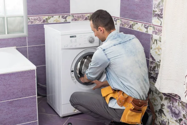 O jovem e bonito reparador em terno de trabalhador com a caixa de ferramentas profissionais está consertando a máquina de lavar roupa no banheiro — Fotografia de Stock