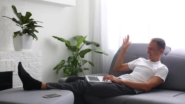 Giovane uomo di bell'aspetto in una camicia jeans nastro sulla tastiera del computer portatile mentre si lavora a casa sul divano. Interni — Video Stock