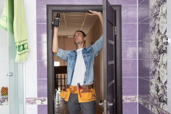 Fiatal ezermester beszerelése egy fehér ajtó elektromos kézi fúró egy szobában. — Stock Fotó